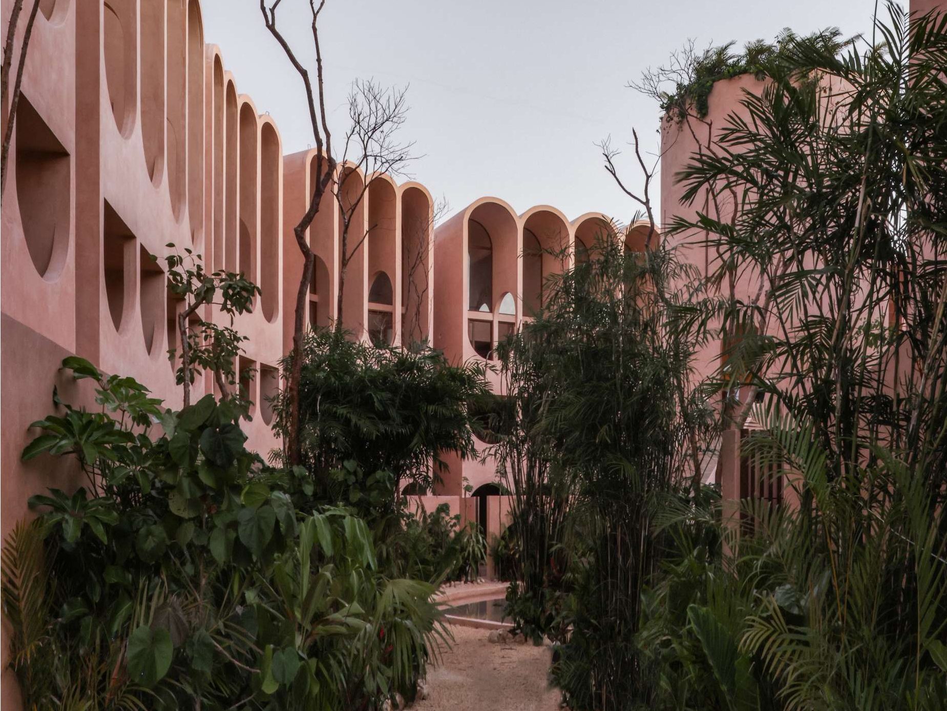 A dreamy pink facade rises behind native landscaping at this Tulum apartment complex, where arched balconies catch the golden light. The curved geometry and minimal palette create a surreal contrast between architecture and the organic jungle surroundings.