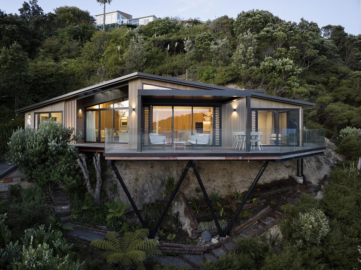 Perched high above the coastline, this striking modern home blends seamlessly into the lush hillside. Cantilevered over the cliff on slender steel supports, it features wraparound glass balustrade decks, warm timber cladding, and floor-to-ceiling glazing that frames panoramic views of the sea and sky. A bold, nature-integrated design that feels like a luxury treehouse.