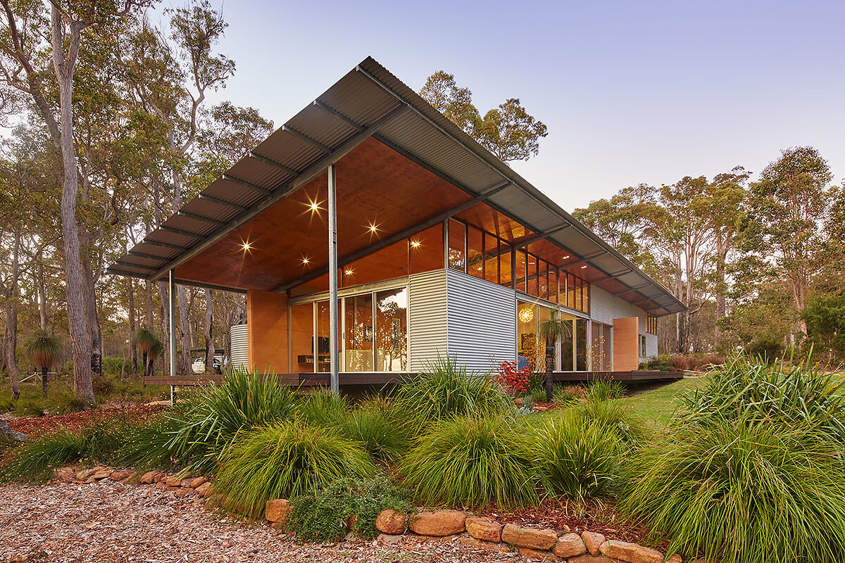 A bushland pavilion with exposed steel frames, rammed earth walls, and a single sweeping roof, inspired by mid-century modern design.